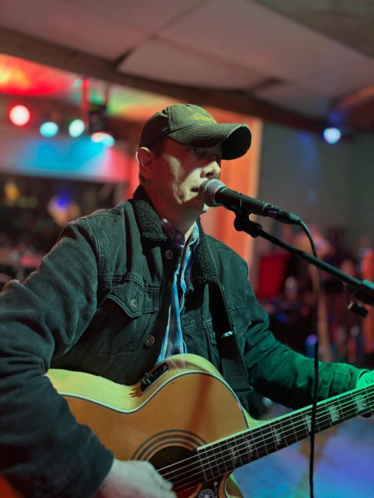 David Habian singing into a microphone with his acoustic guitar on a warmly lit stage